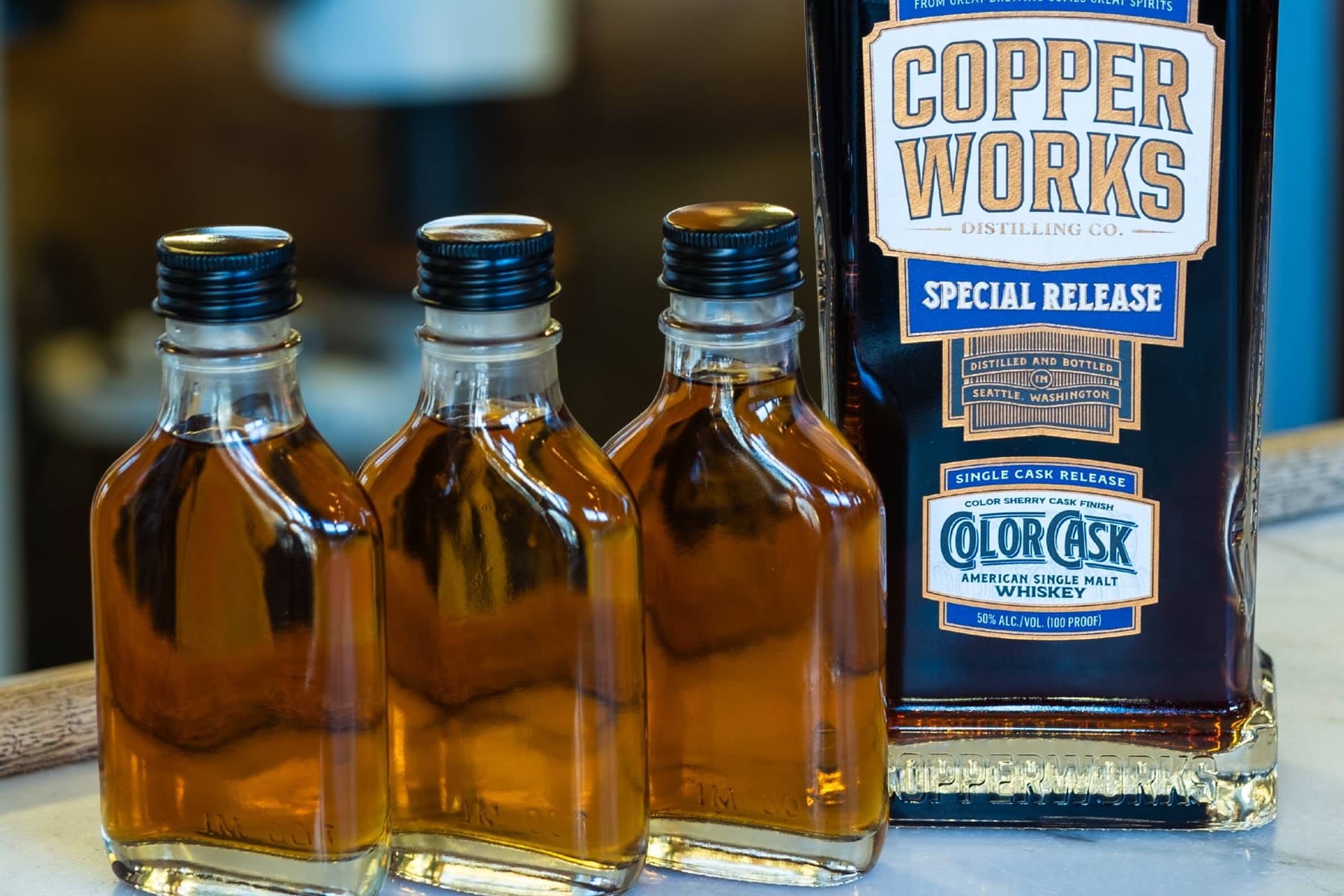 Three 100ml Color Cask Trials sample bottles beside a bottle of Copperworks Color Cask American Single Malt Whiskey on a marble bar.