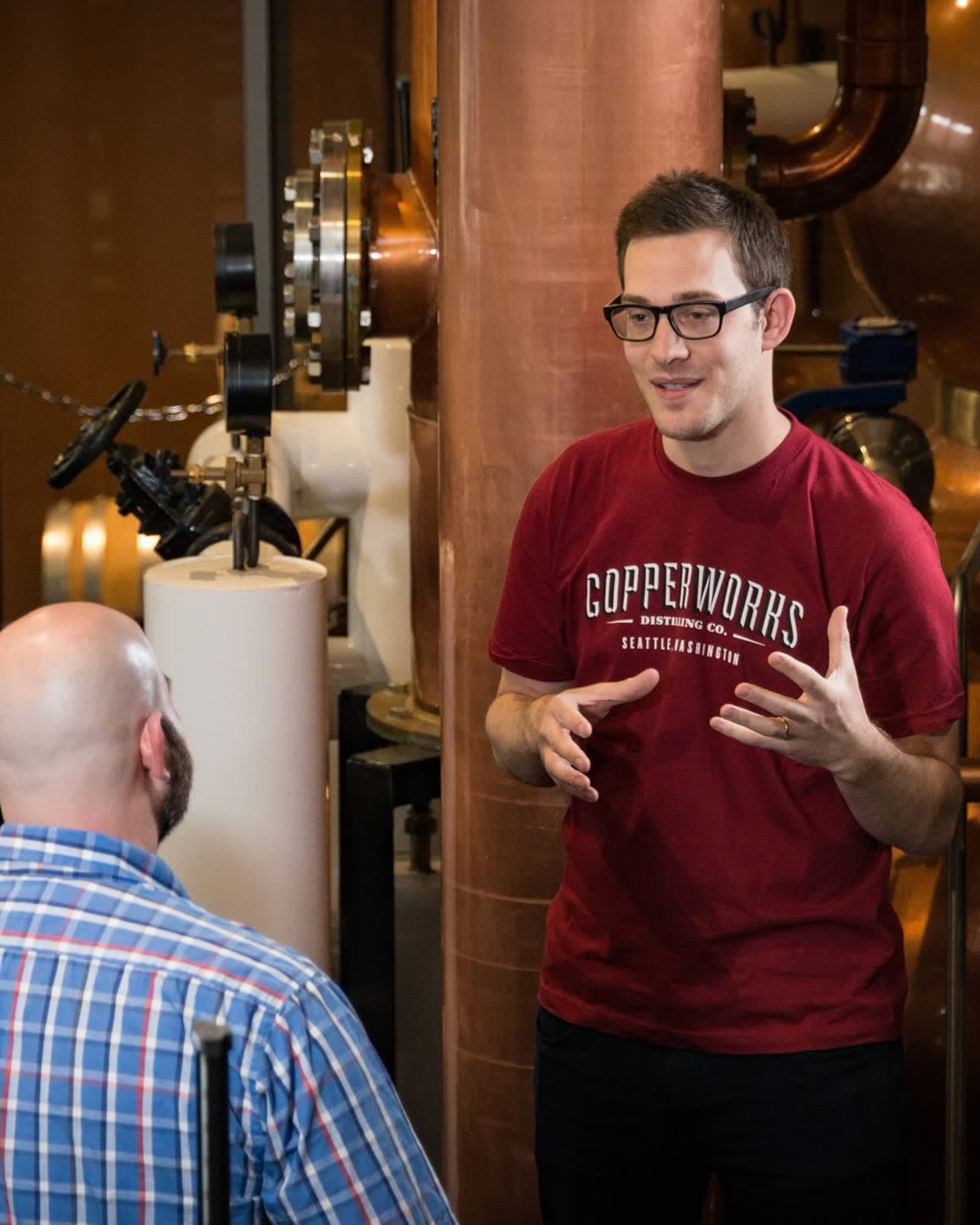 A Copperworks guide talking with a visitor beside the stills during a distillery tour.