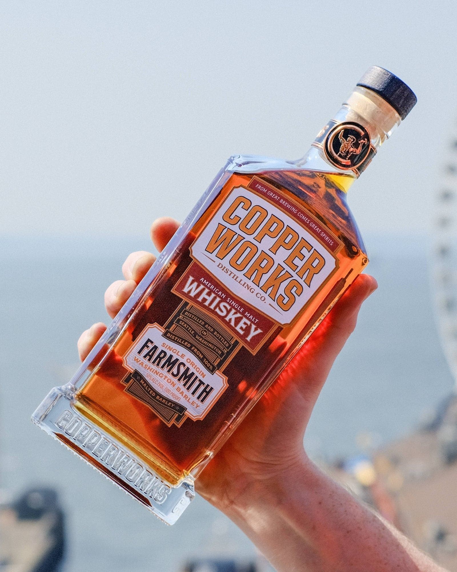 A hand holding up a bottle of Copperworks Farmsmith American Single Malt Whiskey against the sky.