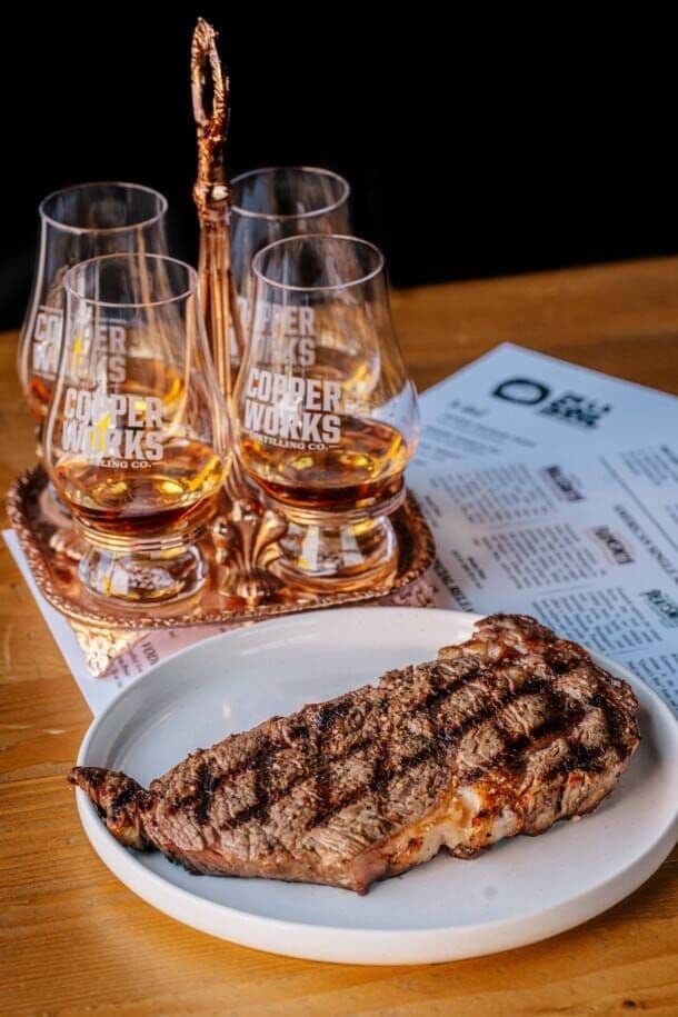 A grilled steak on a plate beside a flight of whiskey in Copperworks tasting glasses.