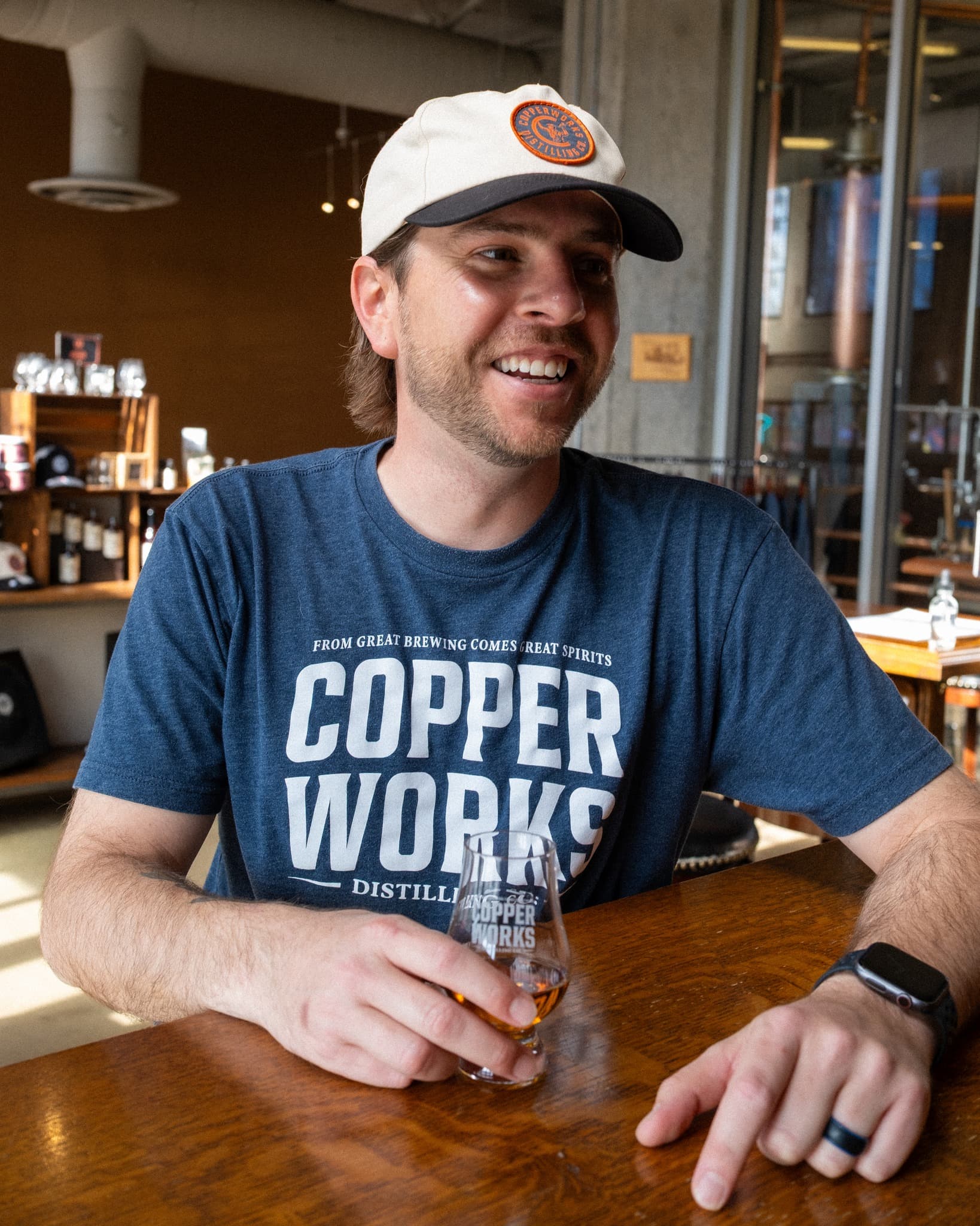 A Copperworks team member at the tasting bar holding a whiskey glass, copper stills visible behind