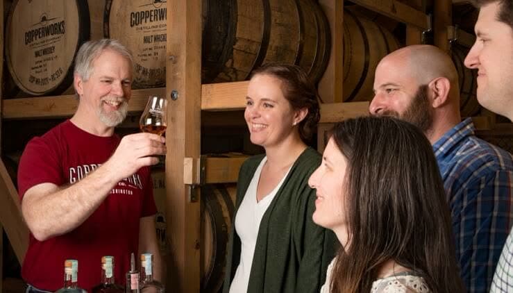 A Copperworks guide raises a glass of whiskey with smiling visitors in the barrel room