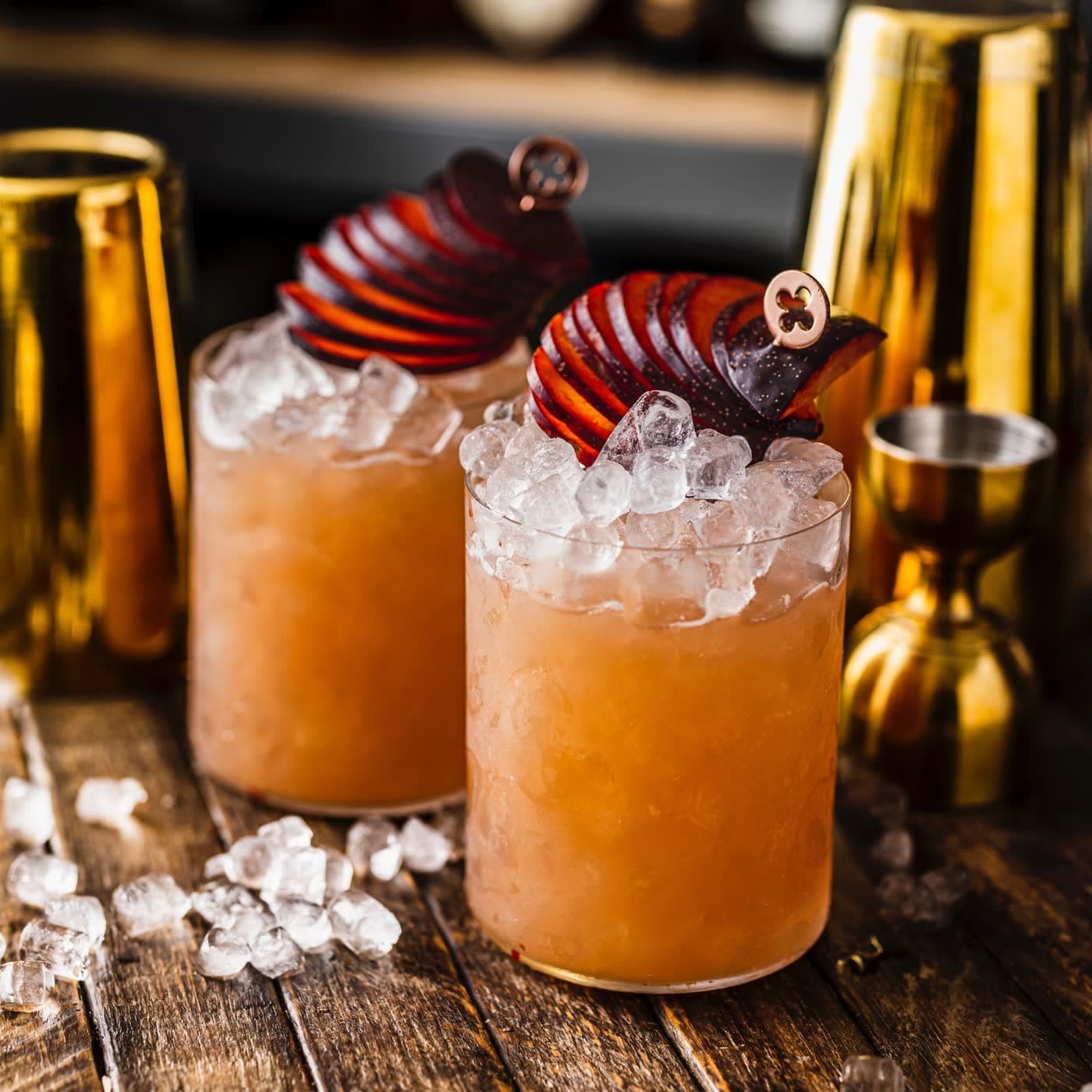 Two Copper Club cocktails over crushed ice, each topped with a fan of sliced plum on a copper pick