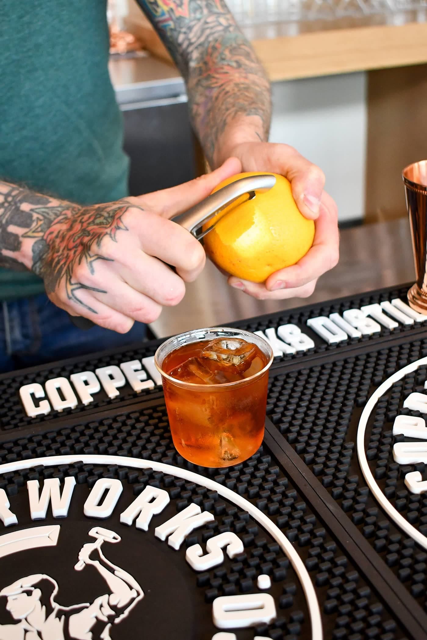 A bartender peels an orange over an Old Fashioned on ice, set on a Copperworks bar mat