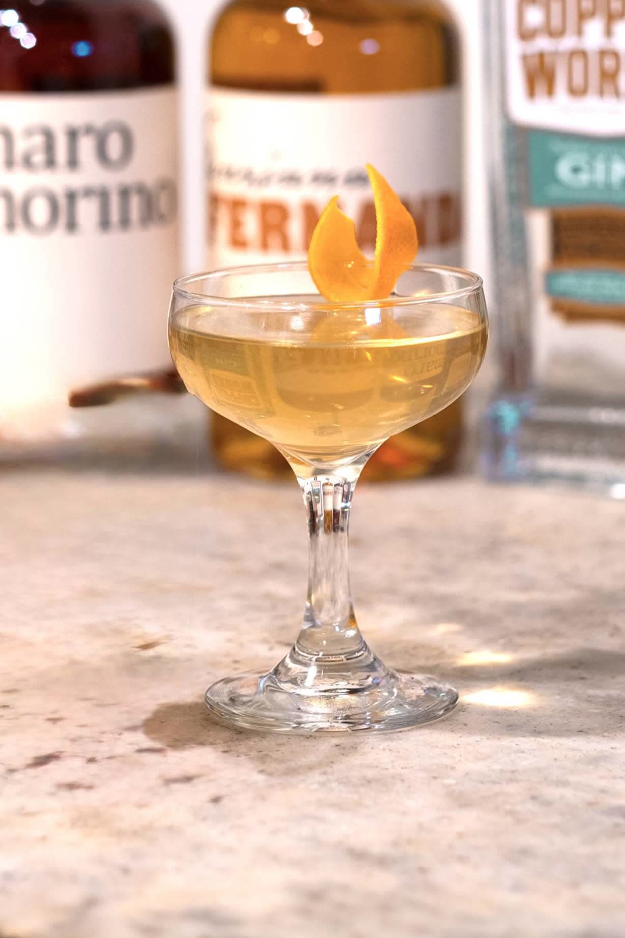 A pale gold White Negroni in a coupe with an orange twist, in front of bottles of Amaro Amorino, Fernanda and Copperworks Gin