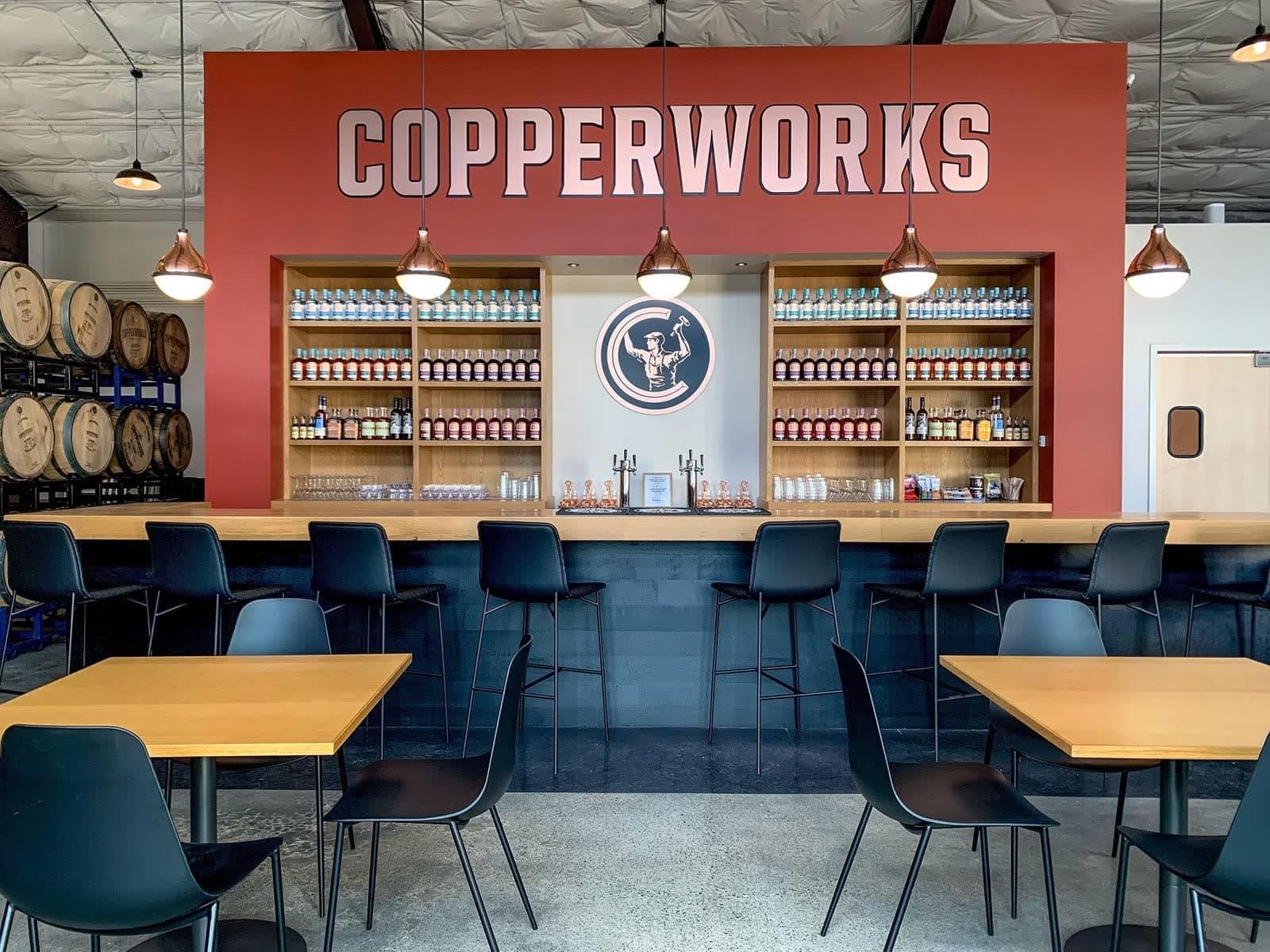 The Copperworks Kenmore bar: a red wall lettered Copperworks, shelves of bottles, copper pendant lights, and barrels stacked to one side