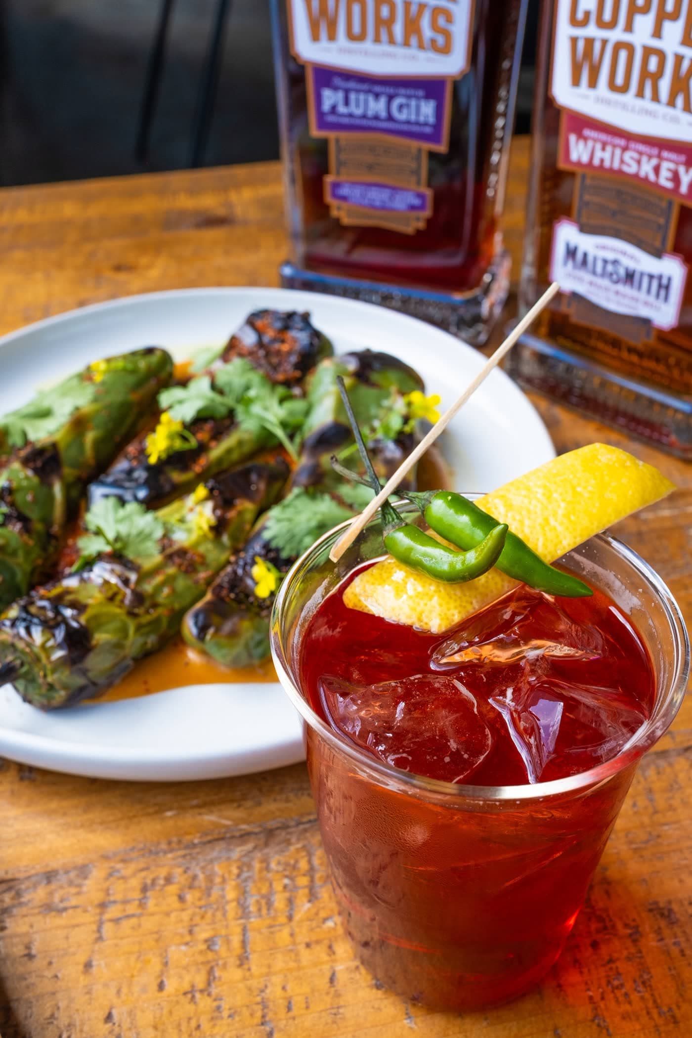 A red cocktail garnished with lemon peel and green chilies beside a plate of charred peppers, with bottles of Copperworks Plum Gin and Maltsmith whiskey behind