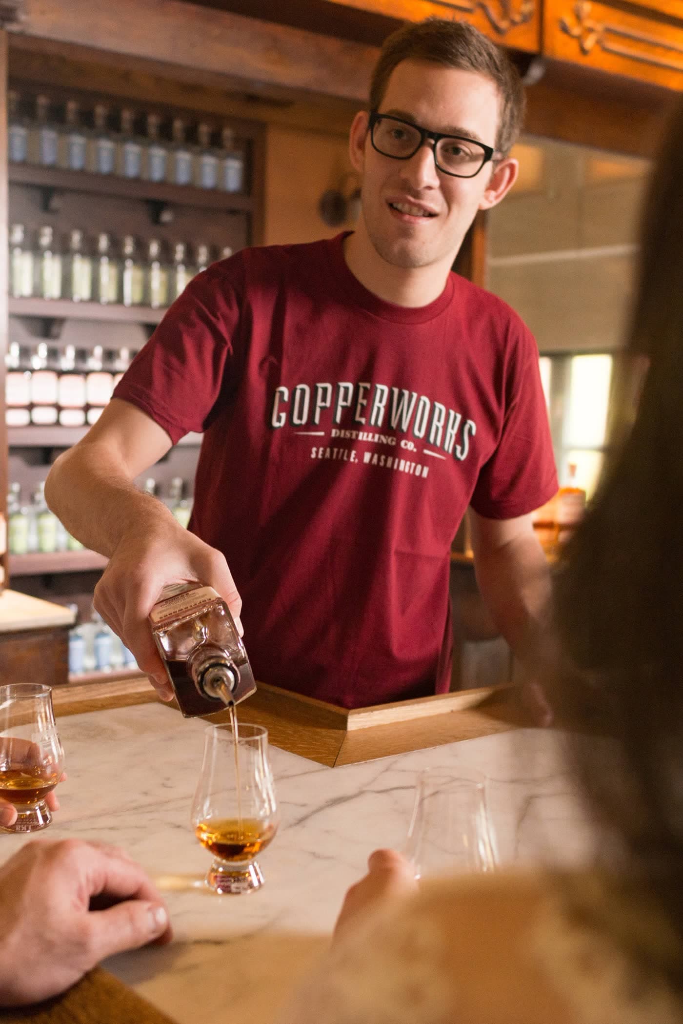 A Copperworks host pours whiskey into a tasting glass across the marble bar of the Seattle tasting room